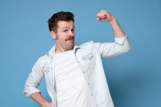 istock Young funny caucasian man in mustache showing his biceps on blue wall. 1200183371