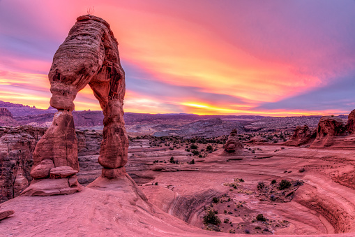 Delicate Arch perched on the edge of a circular ridge against a vibrant sunset sky in Arches National Park, Moab, Utah.