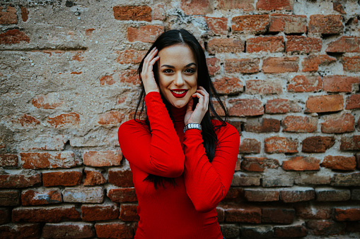 Portrait of a beautiful young woman with long black hair wearing red turtleneck in front of a red brick wall
