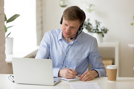 Concentrated pleasant successful male entrepreneur sitting at desk, wearing headset with microphone, watching educational webinar on laptop, writing down notes, head shot. Distant study concept.