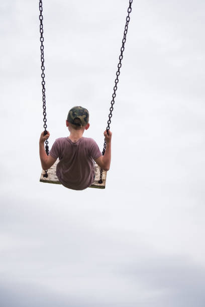 A cheerful boy in a white t-shirt and jeans, standing on a white swing by a serene lake. Ideal for use in advertisements, family blogs, or social media content focused on outdoor activities, childhood