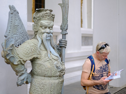 A Western tourist visiting Thailand's famous historical Buddhist Wat Pho temple, studies a guide map next to an old stone-carved Chinese warrior figure statue, standing guard at one of the doors leading into the building that houses the famous reclining Buddha inside the temple complex of the ancient and much visited Buddhist temple of Wat Pho. Interesting relationship between modern tourists and the cultural, historical surroundings of one of Bangkok's famous tourist sites.