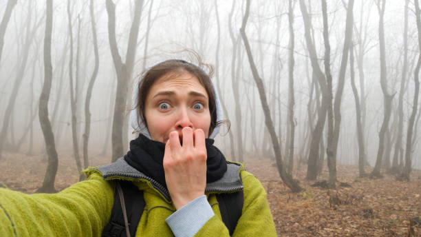 Funny girl traveler while walking through a dangerous foggy forest. Crooked trunks of bare trees in a thick fog. The woman`s hair stands on end. Brunette in fear makes a selfie on the phone. The atmosphere of mystery and fear stock photo