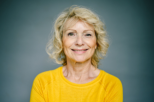 Front view close-up of smiling 59 year old Caucasian woman with medium-length blond hair, pink lipstick, and wearing yellow long sleeved top.