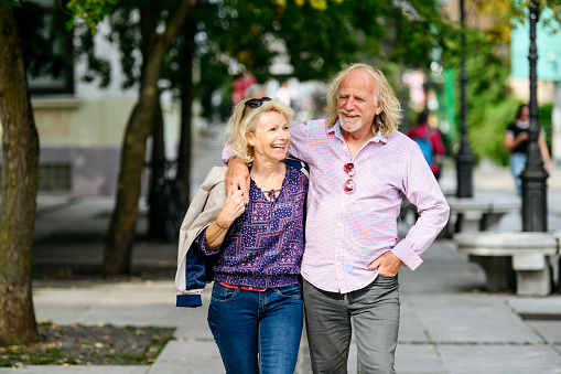 Front view close-up of carefree casual couple in 60s and 70s smiling as they walk with arms around each other in Ljubljana.
