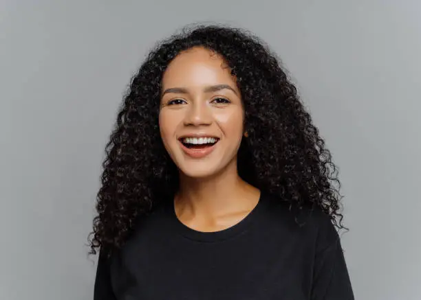 Close up shot of happy dark skinned Afro American woman laughs positively, being in good mood, dressed in black casual clothes, isolated on grey background. Human emotions and feeligs concept.