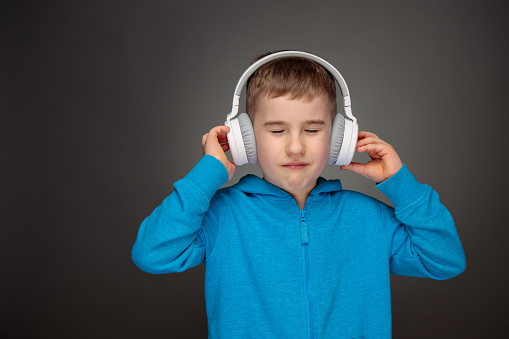 A little boy with white headphones listening music with eyes closed. Studio shot on gray background.