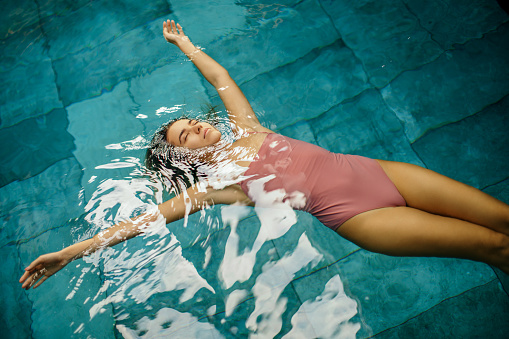 Young woman relaxing and enjoying on health spa poolside. She is floating on water in swimming pool, wears one piece swimsuit and looks beautiful, serene and carefree. Water surface is calm and makes amazing reflection from the window on ceiling