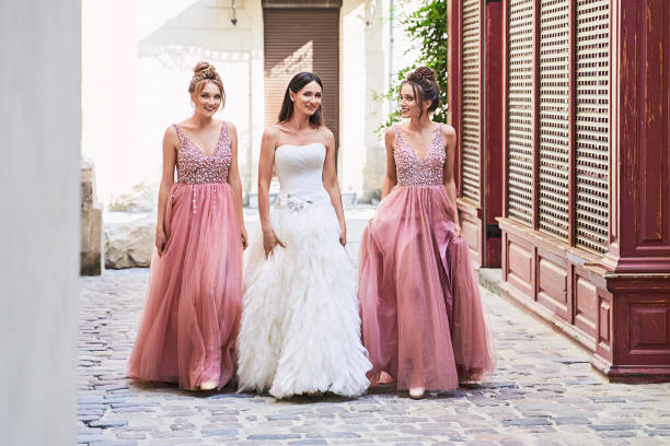 Beautiful bride and bridesmaids walking on the cobblestone street. Maids wear gorgeous elegant stylish red pink violet floor length v neck chiffon gown dress decorated with sequins sparkles and rhinestones holding flowers bouquets. Wedding day. Beautiful bride and bridesmaids in gorgeous elegant stylish red pink violet floor length v neck chiffon gown dress decorated with sequins sparkles and rhinestones holding flowers bouquets. Wedding day in old beautiful European city. pink gown stock pictures, royalty-free photos & images