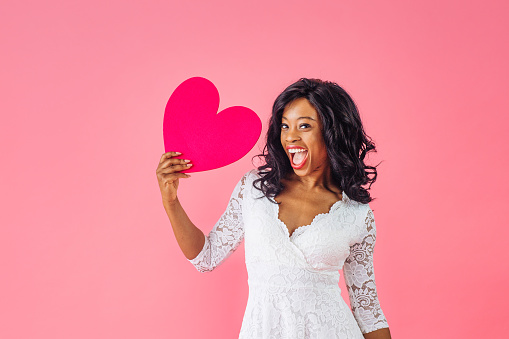 African American woman in late 20s holding heart against pink background.