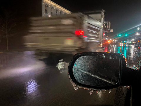 A salt truck is in motion as it passes by. This is a night time shot. The roadway is icy. There is a side view mirror in the bottom of the frame coated with ice.  The scene takes place on a city street. There is copy space in the bottom left of the image