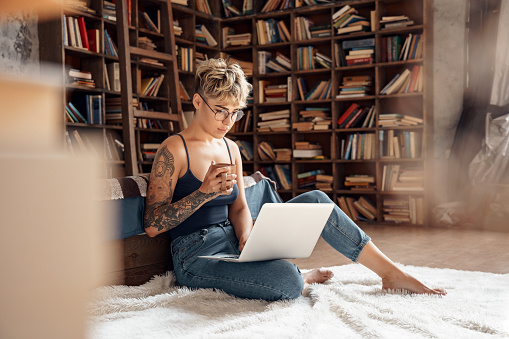 Young woman short hair wearing eyeglasses sitting on fluffy carpet with laptop watching online video lecture distance education holding cup drinking hot coffee relaxed