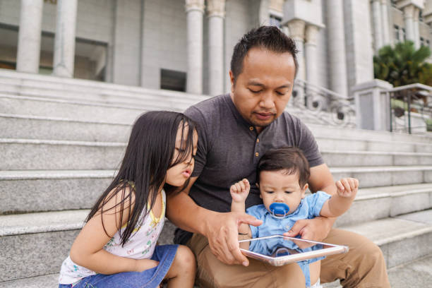 Asian man with his son and daughter using mobile tablet device while sitting down at a public area stock photo