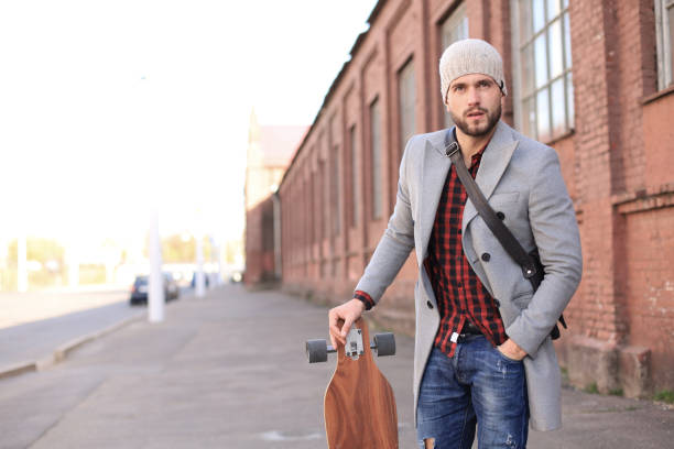 istock Handsome young man in grey coat and hat walking on the street, using longboard. 1200448483