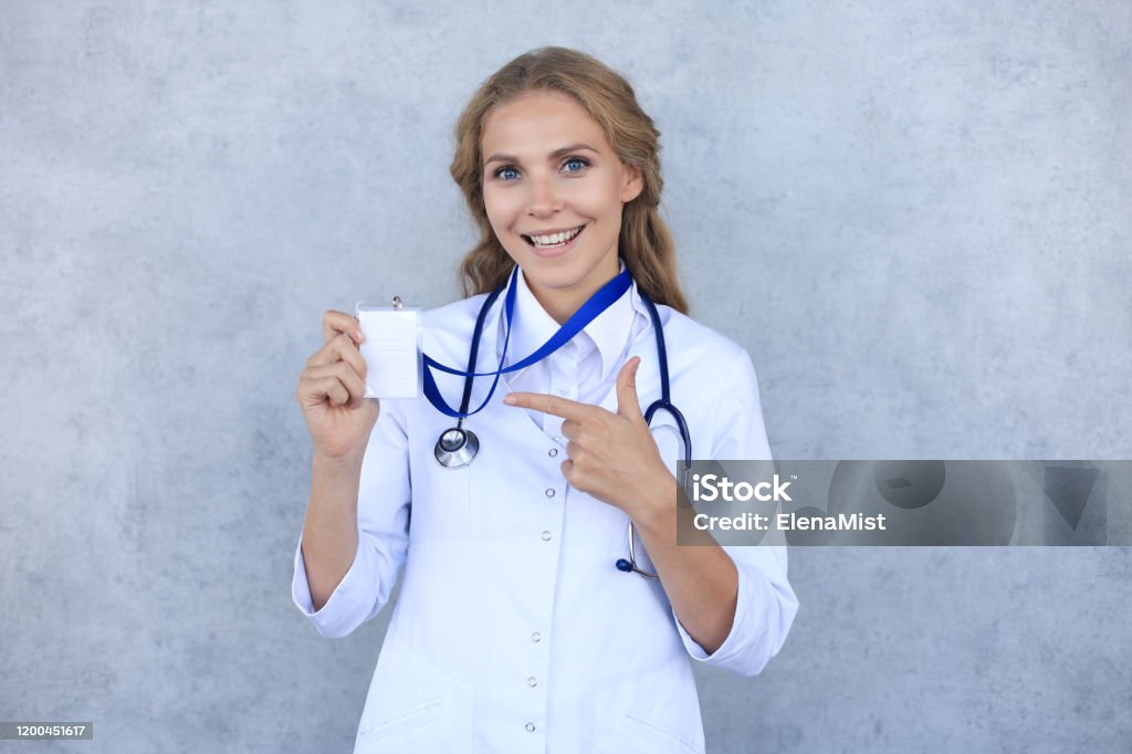 Smiling blonde woman doctor wearing uniform standing isolated over grey background, showing her name on badge. Smiling blonde woman doctor wearing uniform standing isolated over grey background, showing her name on badge Badge Stock Photo