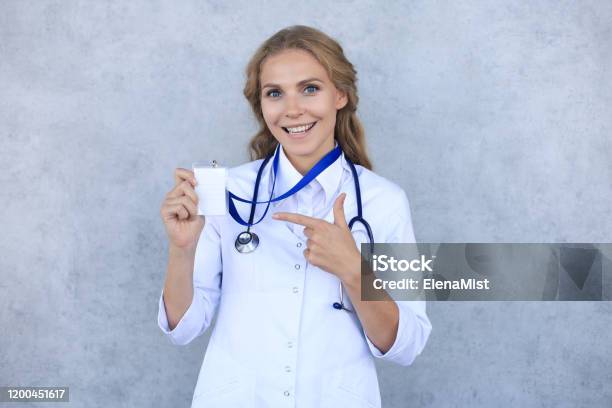 Smiling Blonde Woman Doctor Wearing Uniform Standing Isolated Over Grey Background Showing Her Name On Badge Stock Photo - Download Image Now