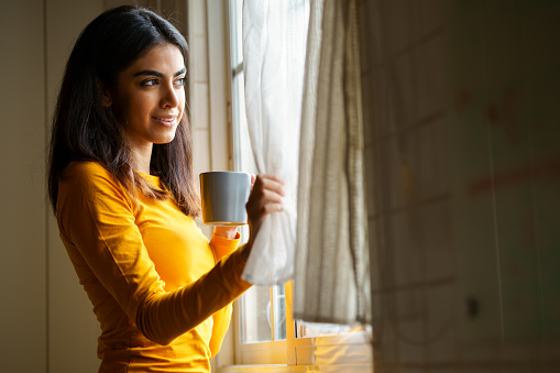 Persian woman drinking coffee at home while looking through the window