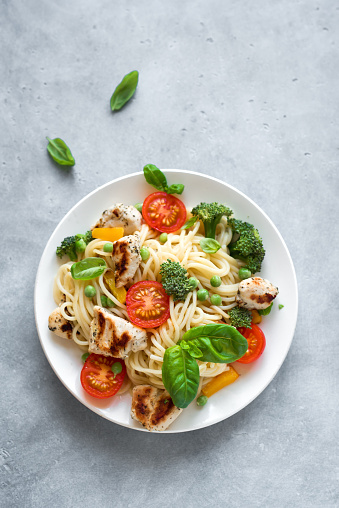 Chicken Pasta Spaghetti Primavera, homemade pasta dish with grilled chicken, vegetables tomato, broccoli, green pea, bell pepper and basil leaves, top view, copy space.