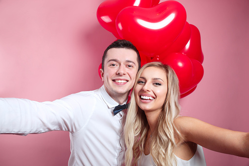 Beautiful young romantic couple taking selfie on mobile phone and standing with red balloons on pink background