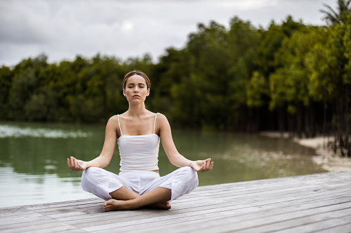 Young woman exercising Yoga in Lotus position on a jetty at river.