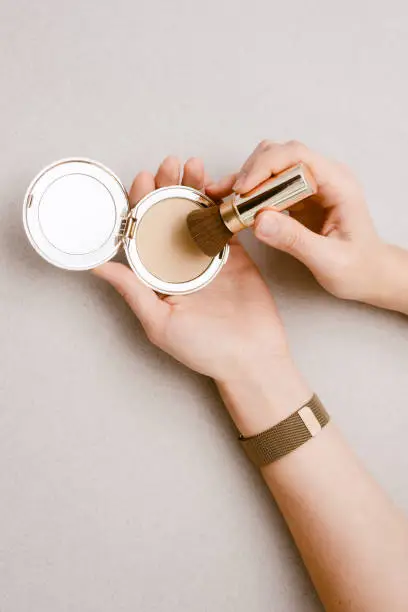 Compact powder with a mirror in the hands of a young girl, close. Pressed tonal base in expensive gold packaging on a beige background. The girl collects powder on a kabuki brush.