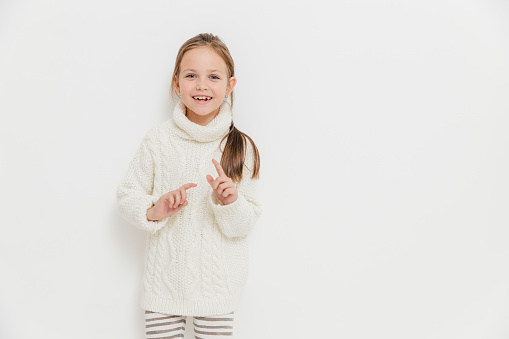 Attractive small female child in warm winter sweater, hears positive story from friend, stands against white background with blank copy space, gestures with hands, has delighted happy expression
