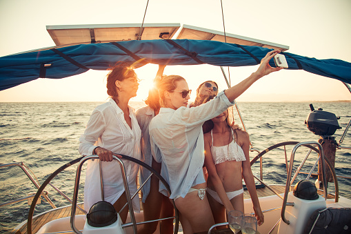 Group of different aged women having a party and making a selfie with mobile phone while sailing on sailboat at sunset. They are very happy and cheerful.