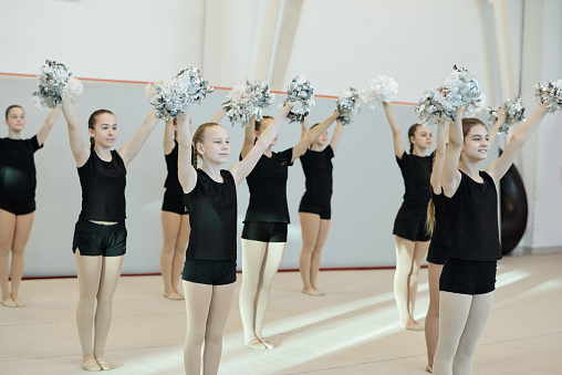 Group of positive school cheerleading girls in black clothing preparing dance with pompoms for competition