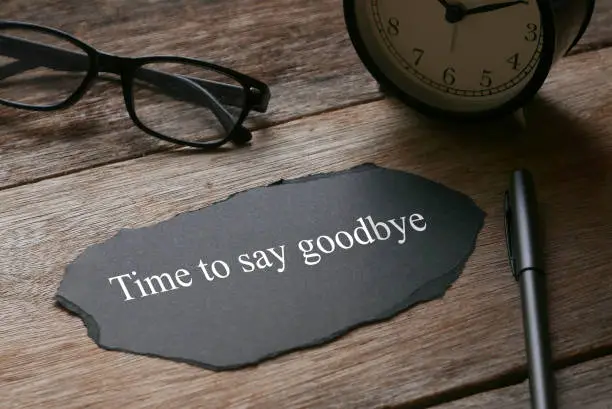 Glasses,clock,pen and a piece of black paper written with Time to say goodbye on wooden background.