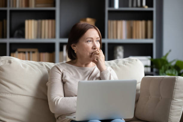 Pensive middle-aged woman look in distance pondering Pensive middle-aged woman sit on couch in living room using laptop look in distance thinking or pondering, thoughtful senior female distracted lost in thoughts feel lonely or sad at home widow stock pictures, royalty-free photos & images