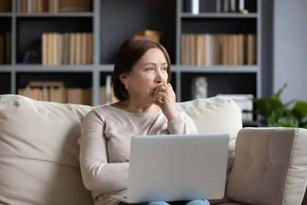 Pensive middle-aged woman sit on couch in living room using laptop look in distance thinking or pondering, thoughtful senior female distracted lost in thoughts feel lonely or sad at home