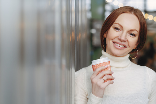 Shot of attractive woman with toothy smile, has makeup, dressed in white turtleneck sweater with collar, holds coffee to go, enjoys spare time, expresses good emotions. People and leisure concept