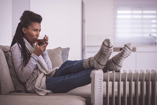 Woman lying on a sofa with woolen socks feet on the radiator heater. The woman drinking hot tea, comfortable sitting on a couch in the living room in the winter season. Using a heater at home in winter.