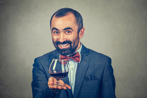 A mature happy man holding showing offering a glass of red wine looking at you camera smiling happily. Mixed race bearded model isolated gray background. Horizontal image. Winemaker waiter concept