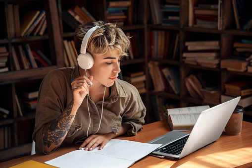 Concentrated and focused young adult woman sitting behind desk with laptop in library, study at university. Student girl writing exercise task and listening music in headset