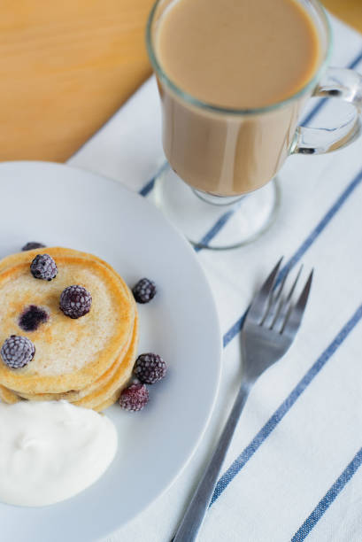 small round fritter or pancakes with blackberry with sour cream on white dish and cocoa on wood table, eastern european breakfast, close up top view of vertical still life stock photo image