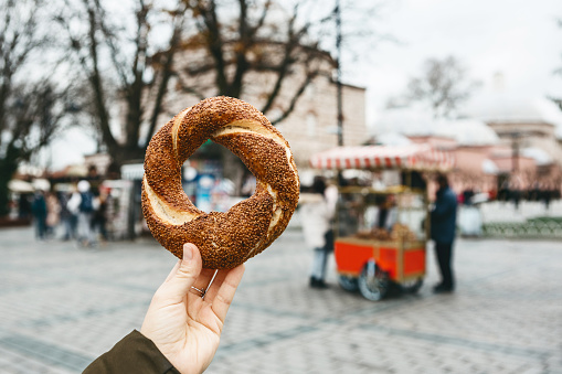 A person is holding a traditional Turkish bagel Simit on the background of a street stall selling Simites. Turkish fast food.
