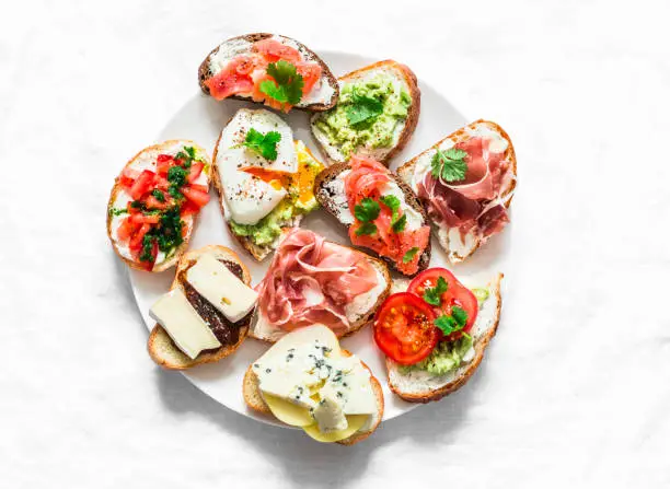 Variety of tapas sandwiches plate - sandwiches with prosciutto, avocado, salmon, egg, tomatoes, jamon, gorgonzola, brie, pear on a light background, top view. Delicious snack, appetizers. Copy space