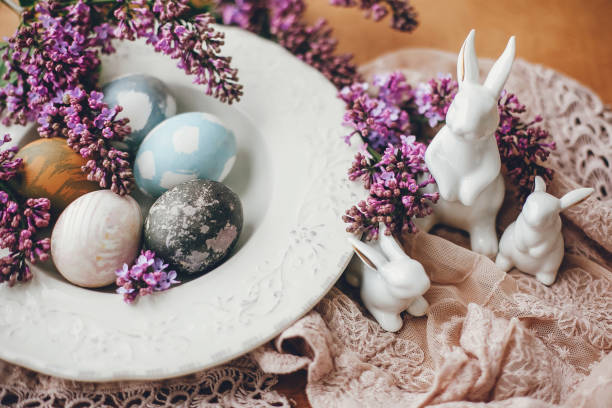 istock Stylish Easter white bunnies, modern easter eggs on vintage plate,  and lilac flowers on rural fabric. Natural dyed easter eggs and spring flowers. Space for text. Holiday decor 1200651514