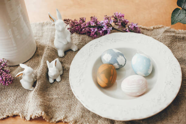istock Stylish Easter eggs on vintage plate, white bunny rabbits and lilac flowers on rustic fabric on wooden table. Rural easter composition of natural dyed eggs and spring purple flowers 1200654055
