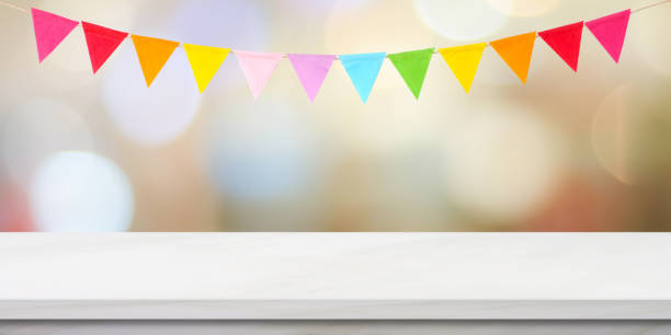 Colorful bunting party flags hanging on blur abstract bokeh light and white marble table background, birthday, greeting card background, banner, backgdrop, mock up, template, wallpaper