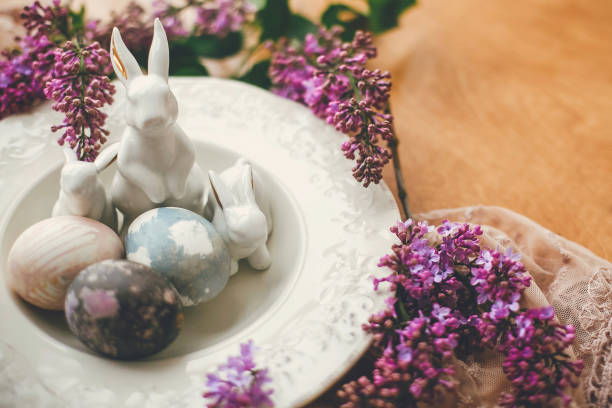 istock Happy Easter. Stylish Easter eggs on vintage plate, white bunnies and lilac flowers on fabric on wooden table. Rural composition of colorful natural dyed easter eggs and spring flowers 1200654940