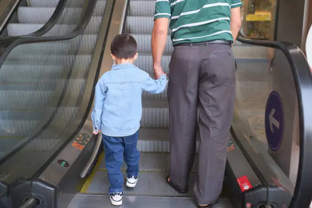 Backside of Little Asian 3 years old toddler boy child and dad holding hands on moving escalator at department store, Father and son spending time shopping, protection concept