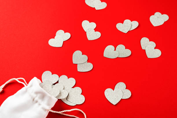 Silver confetti in the shape of heart poured out of white bag on red paper background. Valentine's day concept stock photo