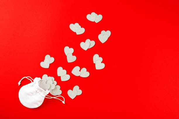 Silver confetti in the shape of heart poured out of white bag on red paper background. Valentine's day concept stock photo