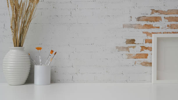 istock Cropped shot of workspace with copy space, paining brush, mock up frame and ceramic vase on white desk with brick wall 1200659966