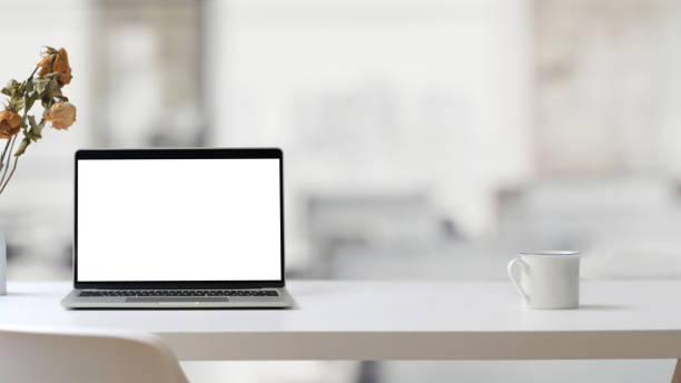 istock Close up view of workplace with open blank screen laptop and coffee cup on white table with blurred office room 1200660542