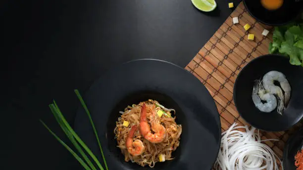 Photo of Overhead shot of Pad Thai, stir fly of Thai noodle with shrimp and egg in black ceramic plate on bamboo placemat with ingredients