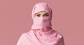 Portrait of young Muslim arabian woman wearing colorful hijab against pink background. Points finger to side. Space for text