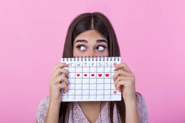 Photo of Portrait of a funny young girl in hiding behind a menstrual periods calendar and looking away at copy space isolated over pink background. Female Period calendar